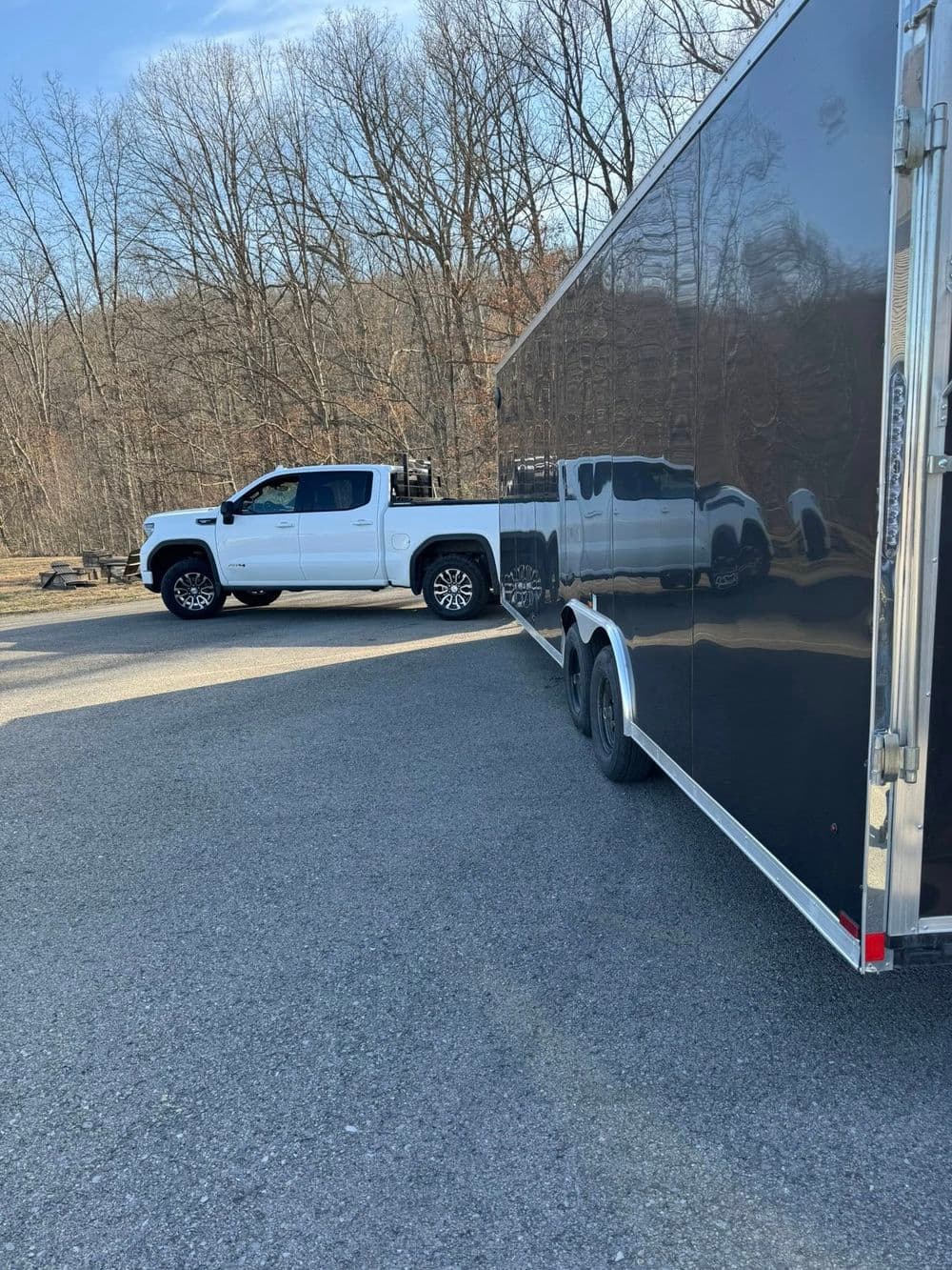 White pickup truck parked next to a black trailer on a gravel surface with trees in the background.
