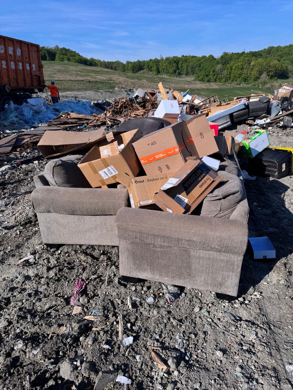 Couch and cardboard boxes piled at a waste disposal site under blue sky and greenery.