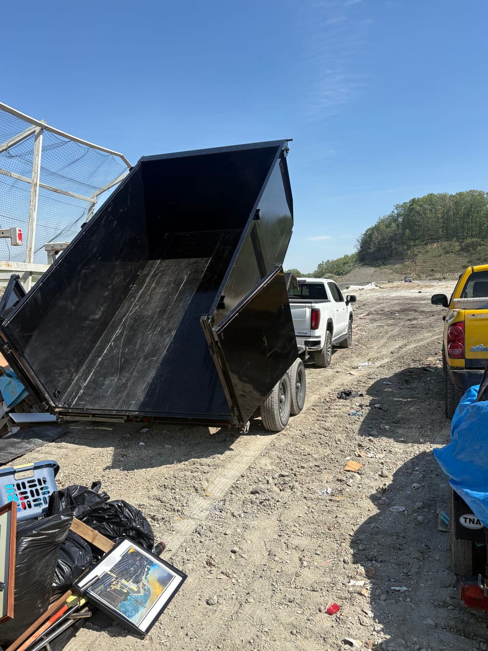 Black dumpster on site with parked trucks and debris in the background under clear blue sky.