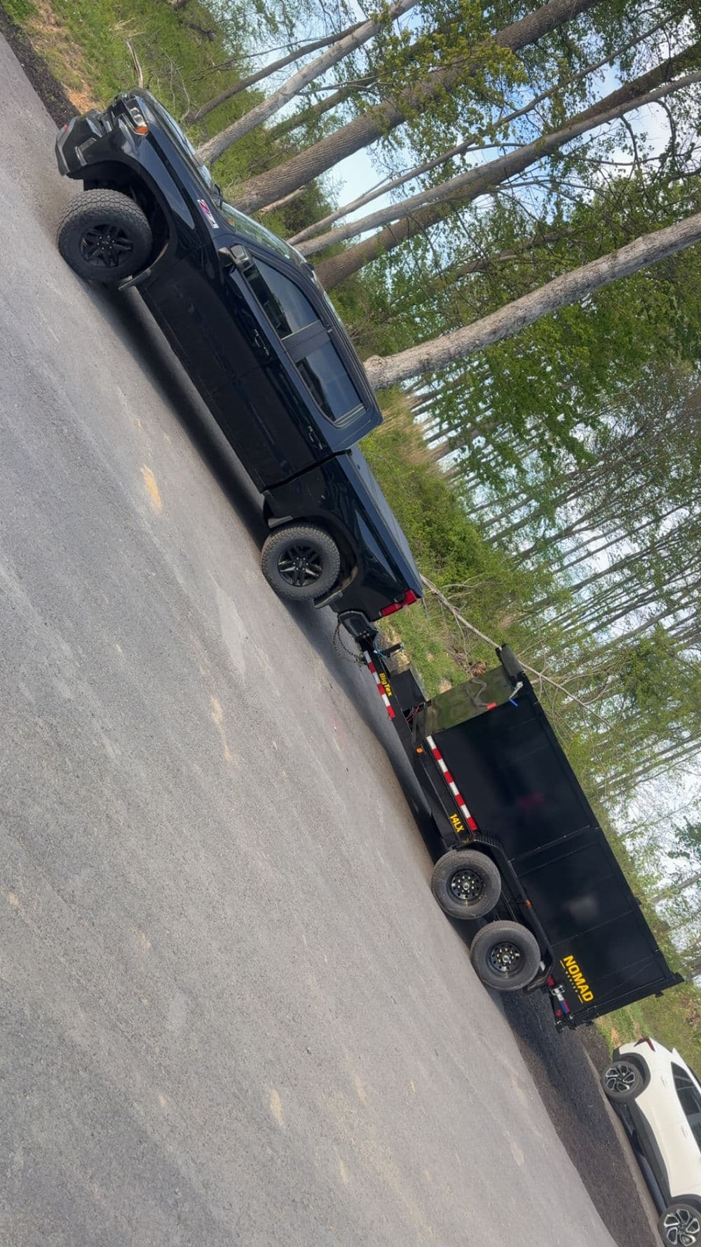 Black pickup truck towing a black dump trailer on a gravel road surrounded by trees.